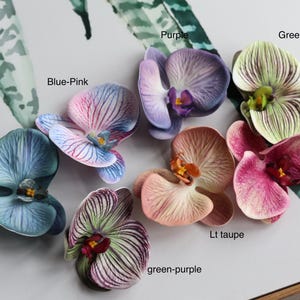 May include: Assortment of ten faux orchid flower heads in various colors, including blue, green, purple, pink, and fuchsia. The flowers are arranged on a white surface.