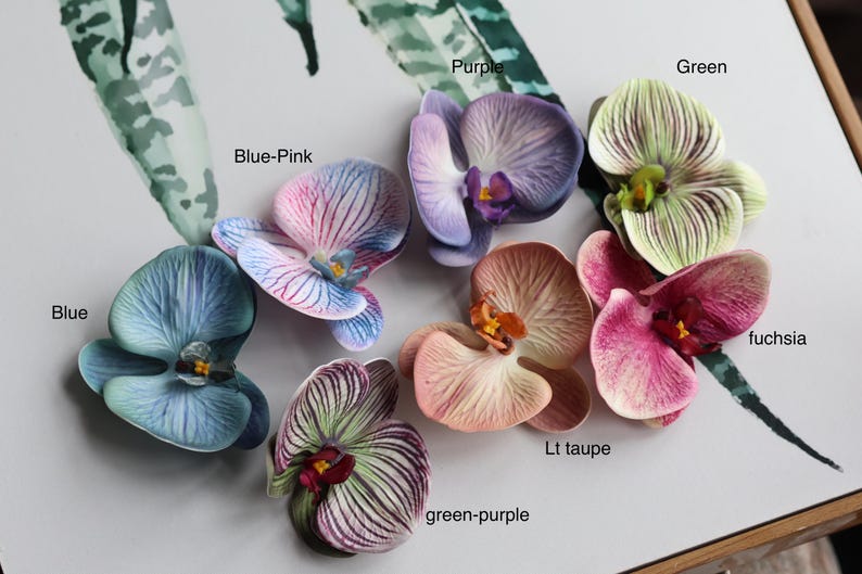May include: Assortment of ten faux orchid flower heads in various colors, including blue, green, purple, pink, and fuchsia. The flowers are arranged on a white surface.