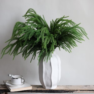 May include: A white ceramic vase with a textured surface holds a bunch of green leafy branches. The vase is sitting on a wooden table.