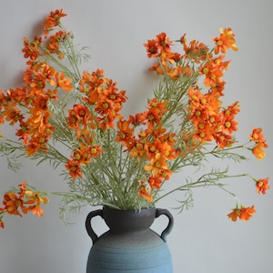 Puede incluir: Un ramo de flores artificiales de color naranja en un jarrón de cerámica azul y marrón. Las flores tienen un aspecto delicado y vaporoso.