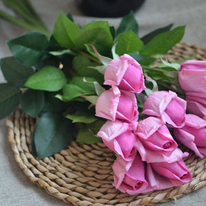 May include: A bouquet of pink artificial roses with green leaves. The roses are arranged on a woven wicker surface.