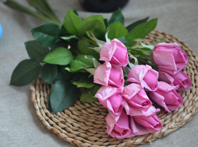 May include: A bouquet of pink artificial roses with green leaves. The roses are arranged on a woven wicker surface.