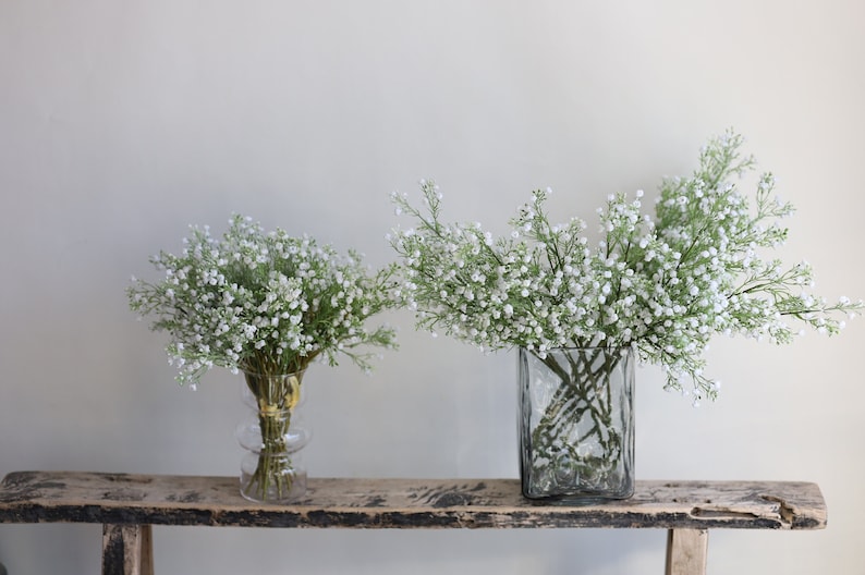 Op de afbeelding: Twee glazen vazen gevuld met witte baby's breath bloemen op een rustiek houten plank.