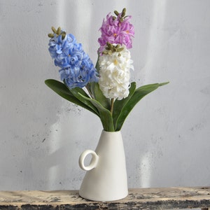 May include: A white ceramic vase with a handle holds three hyacinth flower stems. The flowers are blue, white, and purple, with green leaves. The vase sits on a distressed wooden surface against a light gray wall.