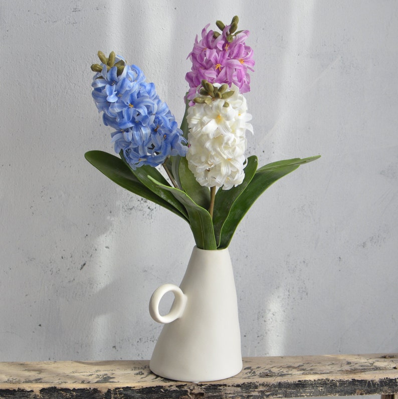 May include: A white ceramic vase with a handle holds three hyacinth flower stems. The flowers are blue, white, and purple, with green leaves. The vase sits on a distressed wooden surface against a light gray wall.