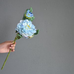 May include: A hand holds a single stem of blue and white artificial flowers with green leaves.
