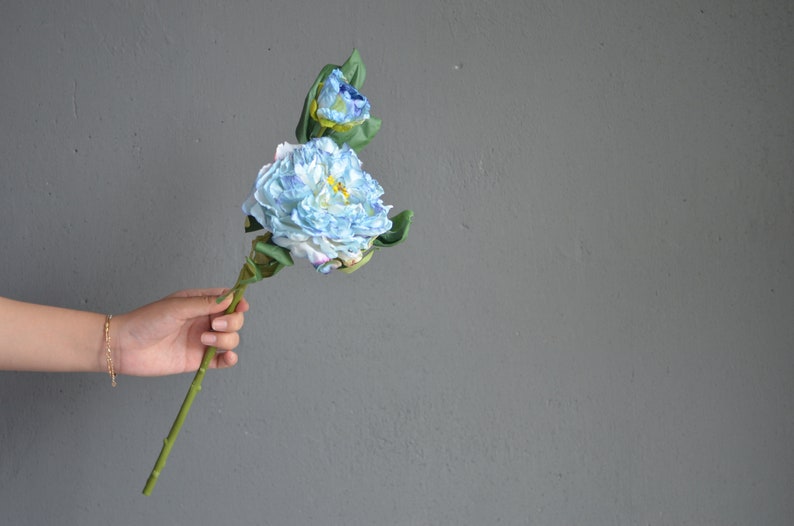May include: A hand holds a single stem of blue and white artificial flowers with green leaves.