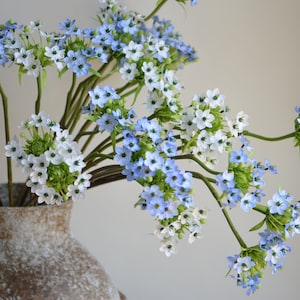 Può includere: Un mazzo di fiori artificiali blu e bianchi in un vaso marrone. I fiori sono disposti a grappolo e hanno un aspetto delicato e arioso.