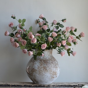 May include: A bouquet of pale pink roses arranged in a rustic, white ceramic vase with a textured surface.