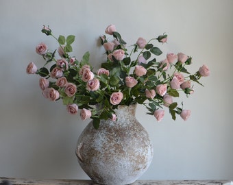 May include: A bouquet of pale pink roses arranged in a rustic, white ceramic vase with a textured surface.