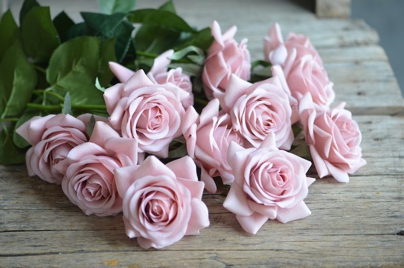 May include: A bouquet of pale pink artificial roses with green leaves on a rustic wooden surface.