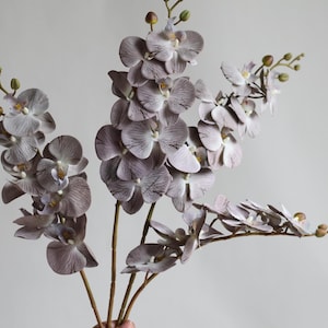 May include: Three stems of artificial orchid flowers with multiple blooms in a light purple color.