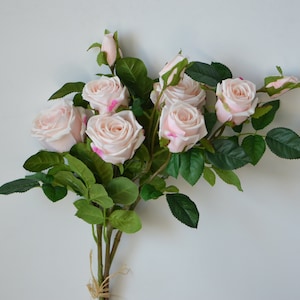 May include: A bouquet of artificial pink roses with green leaves. The roses are a pale pink colour and are arranged in a cluster.