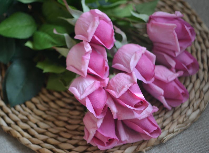 May include: A bouquet of pink artificial roses arranged on a woven wicker surface. The roses are made of fabric and have a realistic appearance.