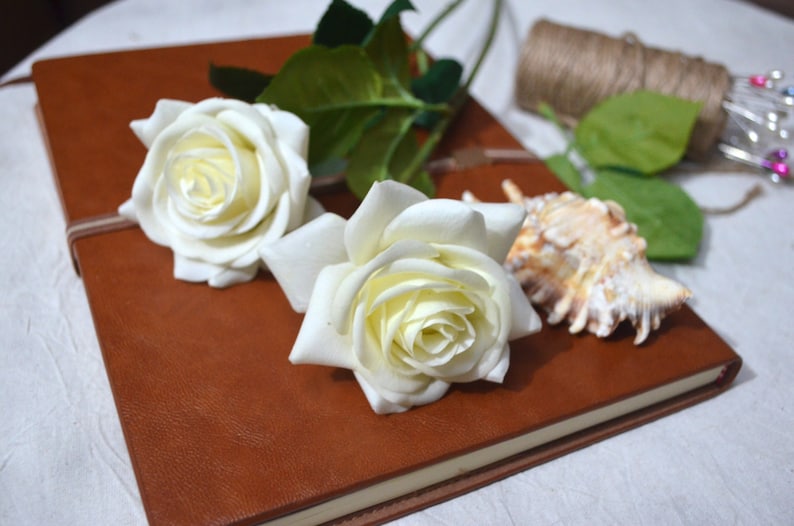 May include: Two white roses with green leaves are placed on top of a brown leather-bound journal.