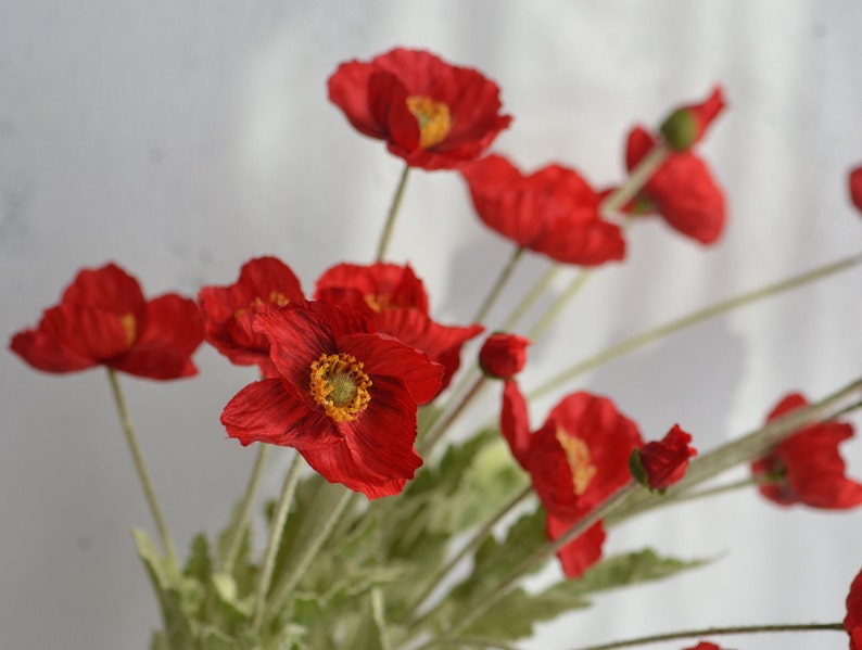 May include: A bouquet of red silk poppy flowers with green stems and leaves. The flowers have a yellow center.