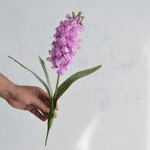 May include: A single artificial hyacinth flower with a stem and green leaves. The flower is a vibrant shade of lavender, with tightly packed florets. The leaves are long and slender. The flower is held against a white background.