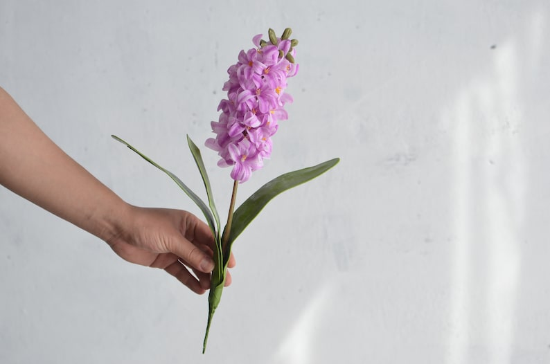 May include: A single artificial hyacinth flower with a stem and green leaves. The flower is a vibrant shade of lavender, with tightly packed florets. The leaves are long and slender. The flower is held against a white background.