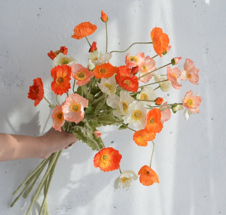 May include: A bouquet of artificial poppy flowers in shades of orange, white, and cream. The flowers have delicate petals and are arranged in a loose, natural style.