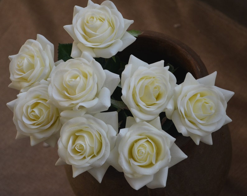 May include: A bouquet of white silk roses arranged in a brown ceramic vase. The roses are arranged in a circular pattern with the stems facing outward.