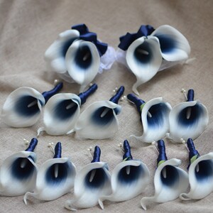 May include: A set of 12 white and navy blue calla lily boutonnieres with navy blue ribbon accents. The boutonnieres are arranged on a beige fabric background.
