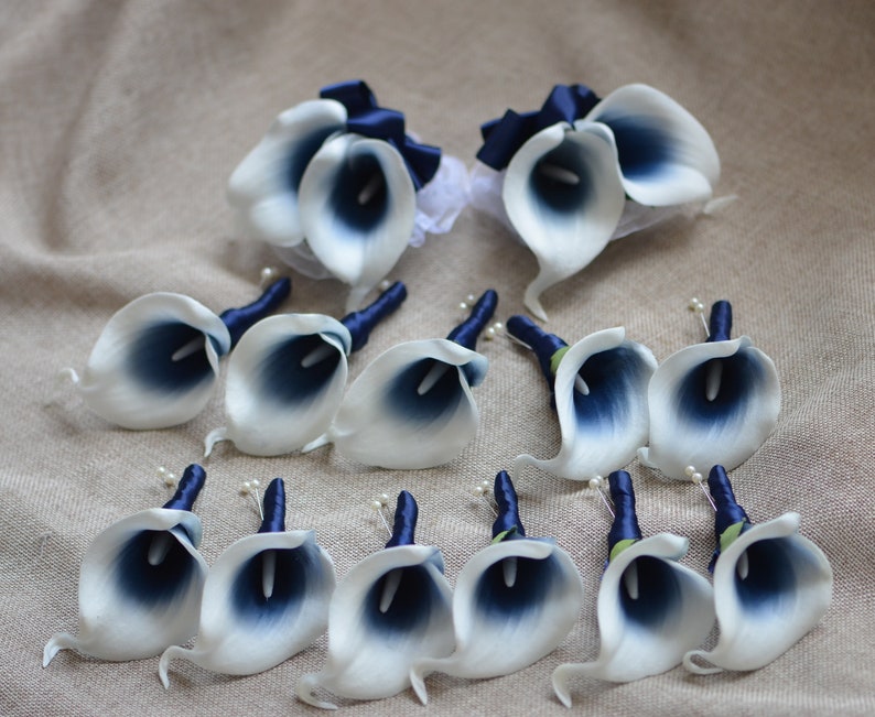 May include: A set of 12 white and navy blue calla lily boutonnieres with navy blue ribbon accents. The boutonnieres are arranged on a beige fabric background.