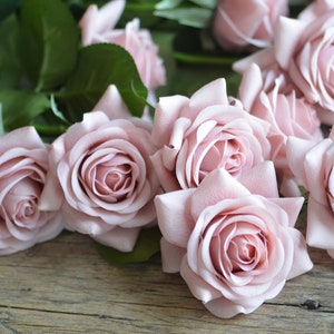 May include: A bouquet of pale pink artificial roses with green stems and leaves. The roses are arranged on a rustic wooden surface.