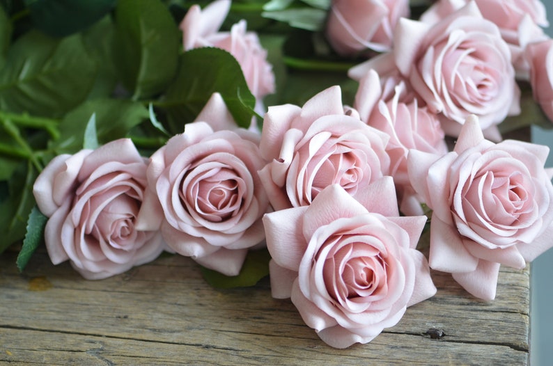 May include: A bouquet of pale pink artificial roses with green stems and leaves. The roses are arranged on a rustic wooden surface.