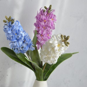 May include: A bouquet of artificial hyacinth flowers in a white vase. The arrangement features three flower stalks in blue, purple, and white, with green leaves. The flowers have a clustered, textured appearance.