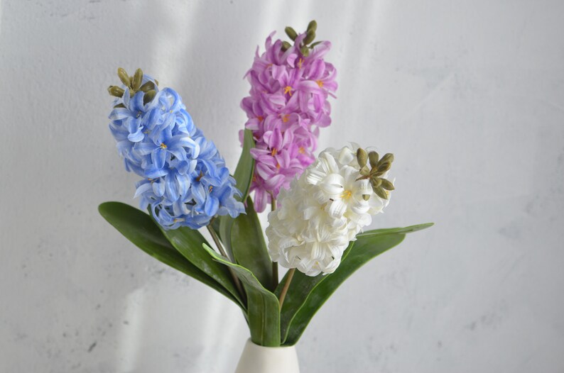 May include: A bouquet of artificial hyacinth flowers in a white vase. The arrangement features three flower stalks in blue, purple, and white, with green leaves. The flowers have a clustered, textured appearance.