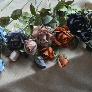 May include: A bouquet of artificial roses in various colors, including pale blue, grey, light brown, burnt orange, black, and dark moss green. The roses are arranged in a row on a textured surface.