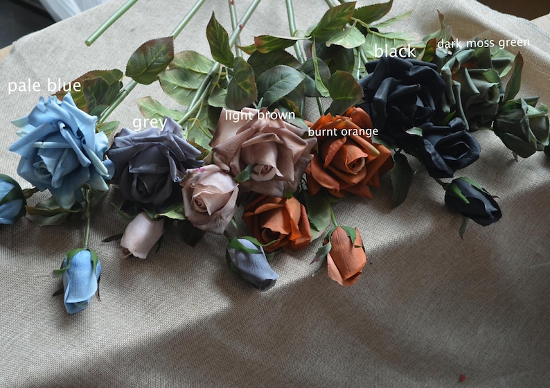 May include: A bouquet of artificial roses in various colors, including pale blue, grey, light brown, burnt orange, black, and dark moss green. The roses are arranged in a row on a textured surface.