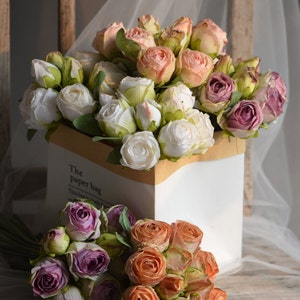 May include: A bouquet of artificial roses in a white ceramic vase with a brown paper bag label that reads "The paper bag". The roses are in shades of pink, white, and peach.