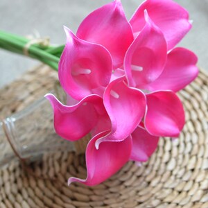 Puede incluir: Un ramo de vibrantes calas rosas con bordes blancos. Las flores est&aacute;n agrupadas, con tallos verdes atados con hilo. El arreglo se encuentra sobre un fondo tejido, con una botella de vidrio transparente visible.