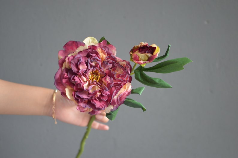 May include: A single stem of artificial peonies with two blooms. The larger bloom is a deep burgundy color with hints of cream and yellow. The smaller bloom is a lighter burgundy color with a yellow center. The stem is green and has leaves.