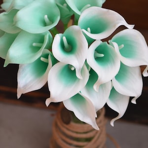 May include: A bouquet of white and teal artificial calla lilies. The flowers are arranged in a vase with a brown rope wrapped around the top.