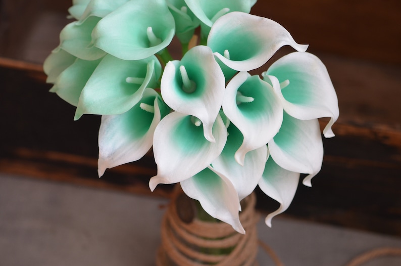 May include: A bouquet of white and teal artificial calla lilies. The flowers are arranged in a vase with a brown rope wrapped around the top.