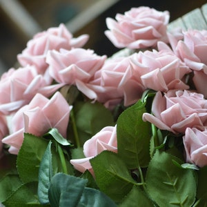 May include: A bouquet of blush pink silk roses with green leaves. The roses are arranged in a cluster, with the stems visible.