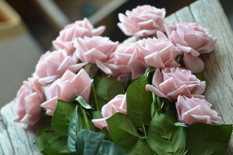 May include: A bouquet of blush pink silk roses with green leaves. The roses are arranged in a cluster, with the stems visible.