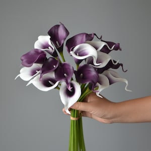 Puede incluir: Un ramo de lirios calla artificiales con pétalos blancos y morados. Los tallos son verdes y están atados con una cinta marrón.