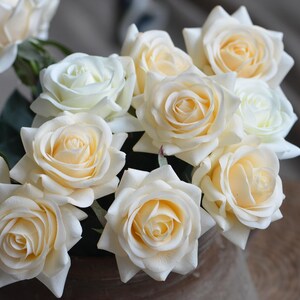 May include: A bouquet of cream-colored artificial roses with a soft, velvety texture. The roses are arranged in a vase and have a natural, realistic appearance.