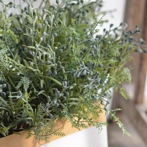 Può includere: Un mazzo di verde artificiale verde e grigio in un vaso marrone. Il verde ha piccoli boccioli grigi rotondi.