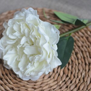 May include: A single white artificial peony flower with green leaves, arranged on a woven brown wicker surface.