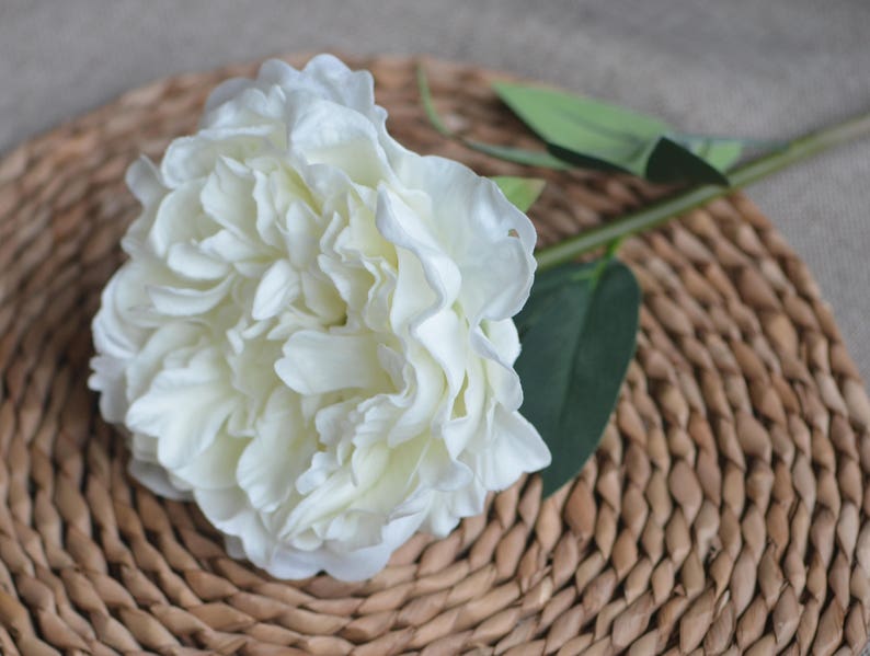 May include: A single white artificial peony flower with green leaves, arranged on a woven brown wicker surface.