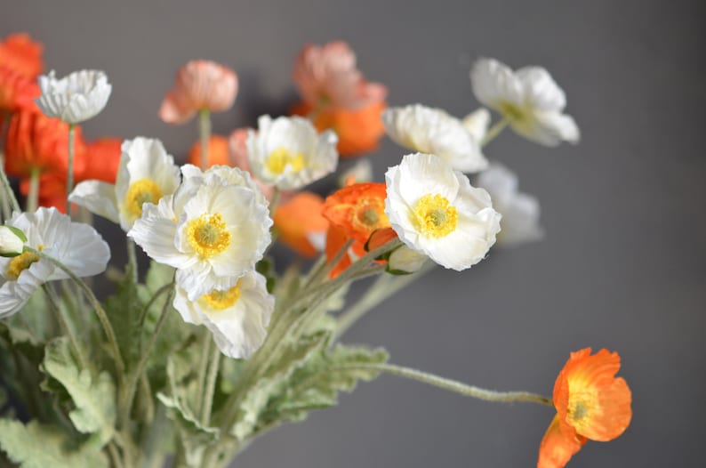 Puede incluir: Un ramo de flores artificiales con flores de amapola blancas y naranjas. Las flores tienen un aspecto realista y est&aacute;n dispuestas en un jarr&oacute;n.