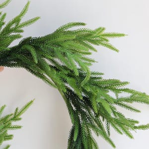 May include: A green artificial pine garland with long, thin branches. The garland is arranged in a circle, creating a wreath shape.