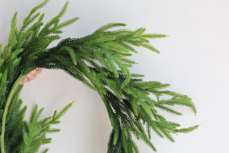 May include: A green artificial pine garland with long, thin branches. The garland is arranged in a circle, creating a wreath shape.