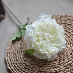 May include: A single white artificial peony flower with green leaves on a woven brown wicker surface.