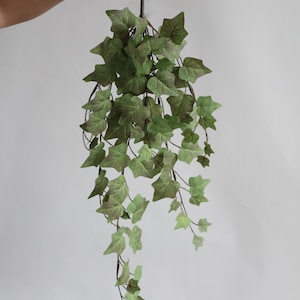 May include: A hanging artificial ivy plant with green and brown leaves. The leaves are shaped like stars and are attached to thin, dark stems. The plant is held up by a hand against a plain white background. This decorative plant is suitable for home decor.