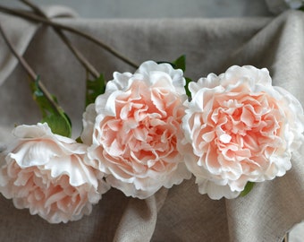 May include: Three artificial peach-colored peonies with white edges on a light brown linen background.